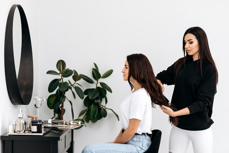 Beauty professional smiling while styling a seated client's long hair in a bright, quiet studio, showing how Sandy Springs salon suites help protect a stylist's energy.